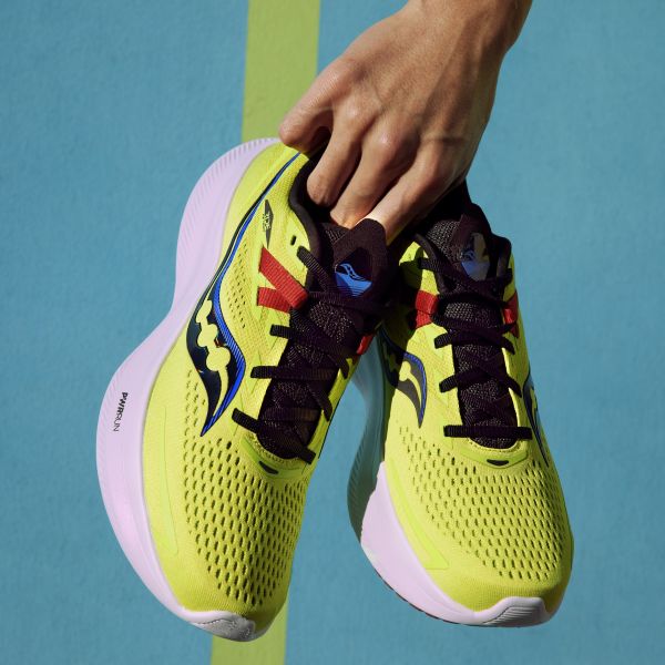 Eine Hand hält ein Paar neon-gelbe Saucony Ride 15 Laufschuhe vor blauem Hintergrund.