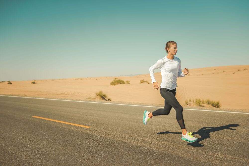 Eine Läuferin in weißem Langarmshirt, schwarzer Tight und Saucony Laufschuhen joggt auf einer leeren Asphaltstraße durch eine sonnige Wüstenlandschaft mit Sanddünen.
