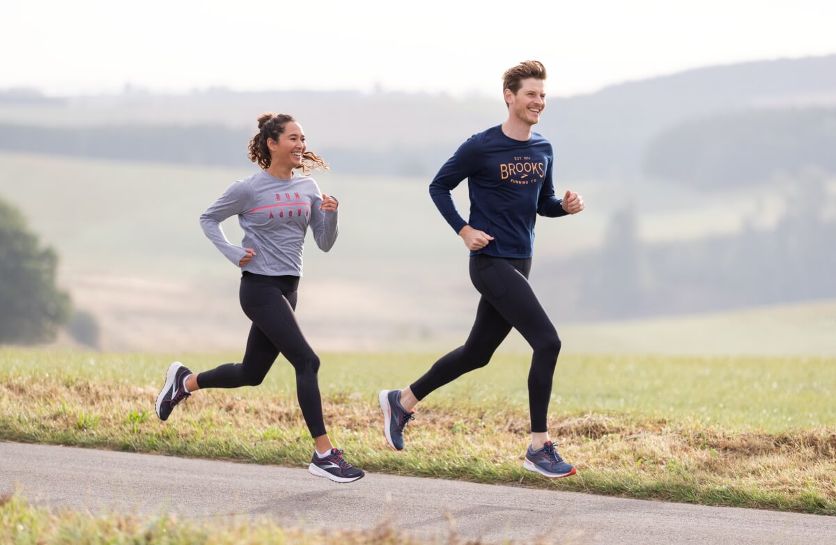 Ein lächelndes Paar beim gemeinsamen Joggen auf einer asphaltierten Landstraße in einer hügeligen Landschaft.