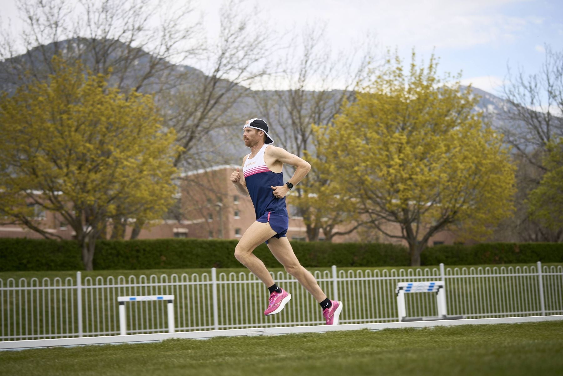 Ein männlicher Läufer in blauer Sportbekleidung und pinken Laufschuhen joggt konzentriert auf einer Tartanbahn vor einer Kulisse aus Bäumen und Bergen.