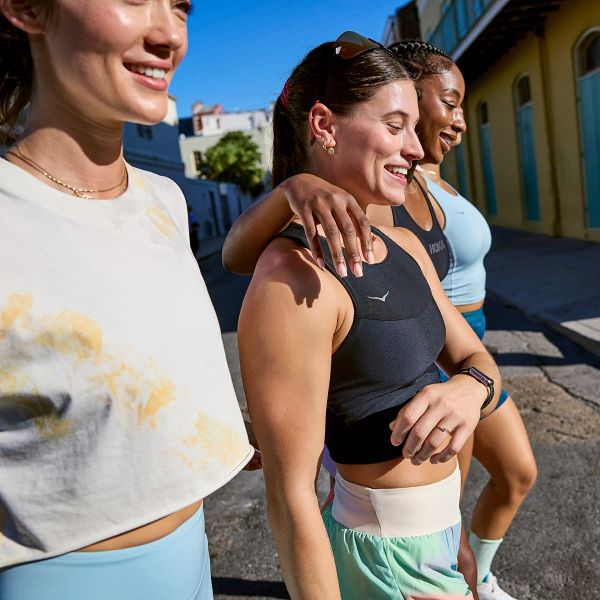 Drei lachende Frauen in sportlicher HOKA-Laufbekleidung spazieren gemeinsam eine sonnige Stadtstraße entlang.