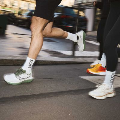 Mehrere Läufer in Bewegung auf Asphalt mit verschiedenen Saucony Laufschuh-Modellen, Fokus auf dem weißen Modell mit grüner Sohle.