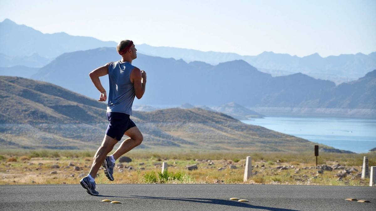 Ein männlicher Läufer in grauem Tanktop und dunklen Shorts joggt auf einer Straße vor einer weiten Berglandschaft und einem blauen See.